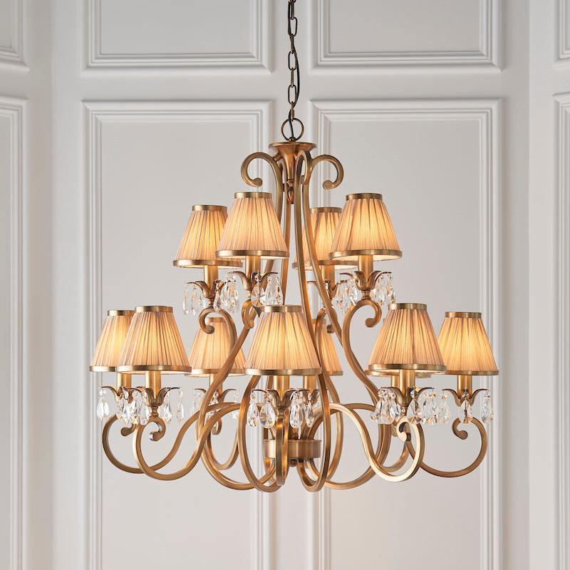Interiors 1900 Oksana 12-Light Chandelier in Antique Brass with Beige Shades – A classic chandelier featuring antique brass detailing, faceted crystal accents, and beige fabric shades. Adjustable height (78cm-177cm), dimmable compatibility, and uses 12 x E14 bulbs (not included).